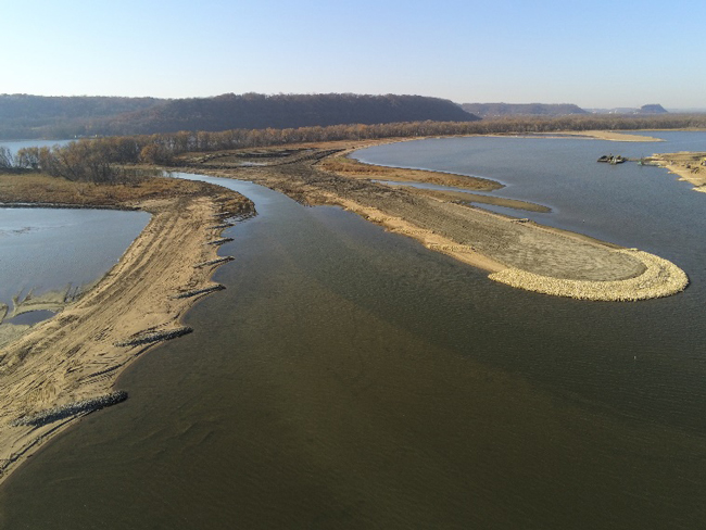 Aerial view of a river restoration project under construction, showing newly formed sandy and rock-lined islands and channels curving through the water, with wooded bluffs in the distance.
